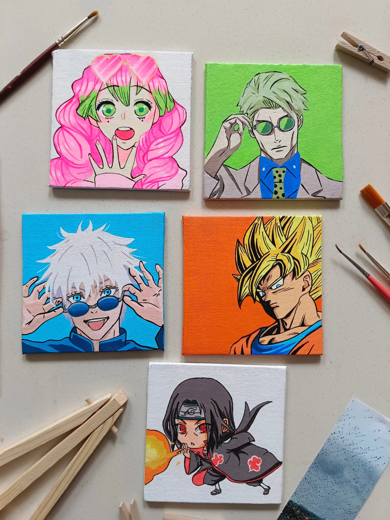 Colourful set of acrylic mini canvases featuring Mitsuri Kanroji, Sanji, Satoru Gojo, Son Goku and Itachi Uchiha, arranged with brushes and clips.