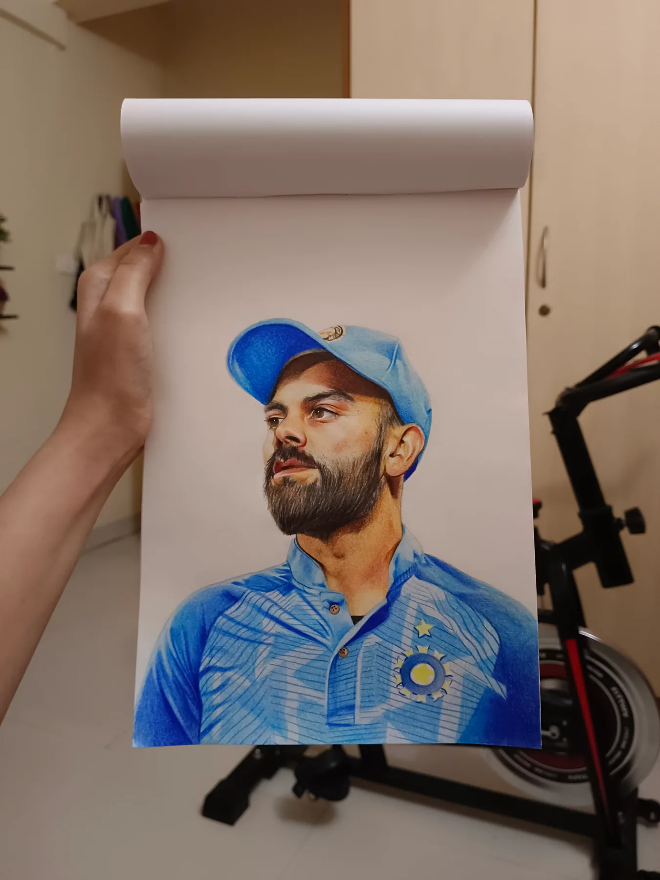 Colour-pencil portrait of Virat Kohli from the Indian cricket team in a blue jersey, shown on a sketchpad as a custom sports artwork.