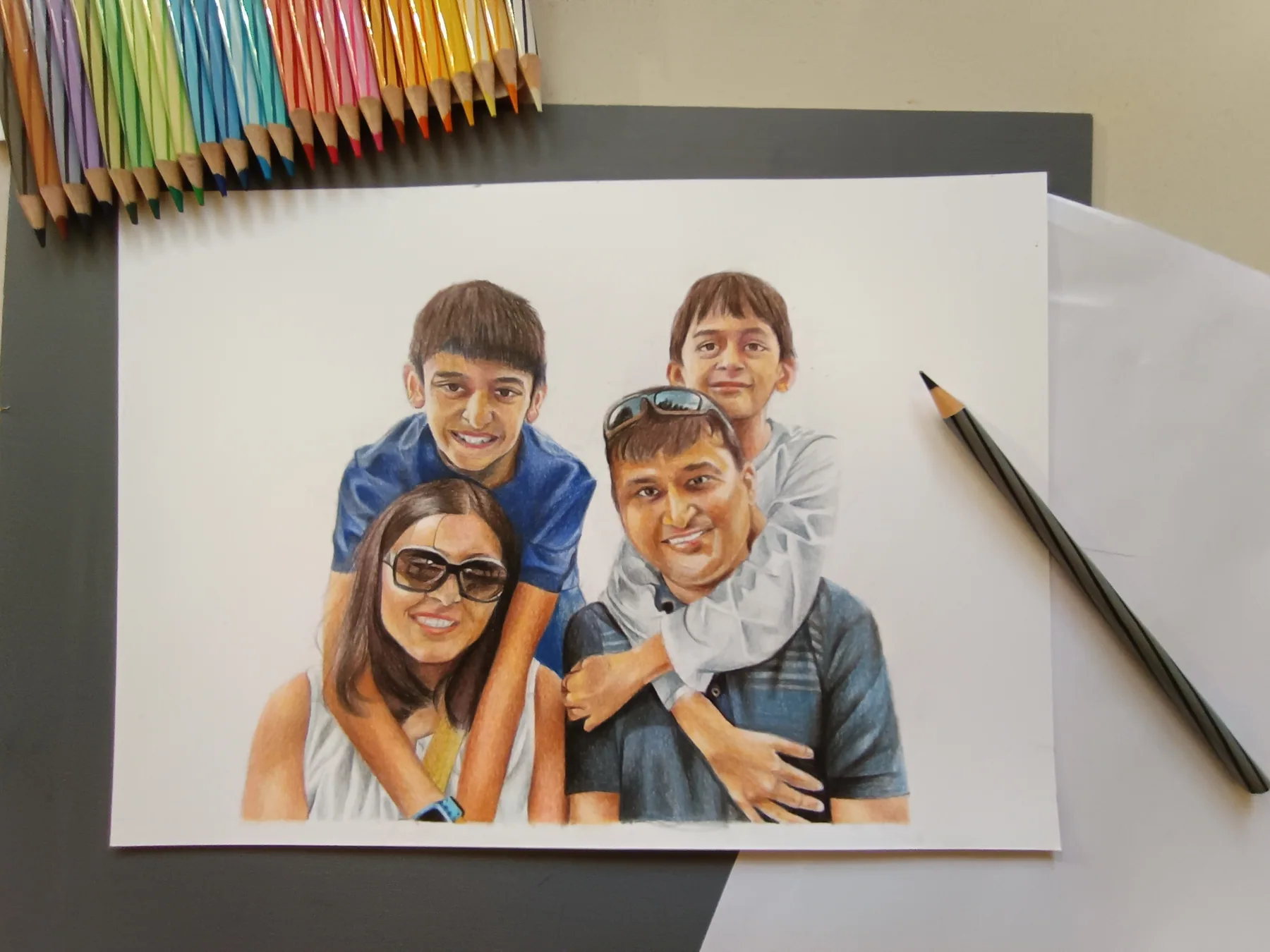 Colour pencil family portrait from a photo, shown on paper with pencils nearby as a custom commission example.